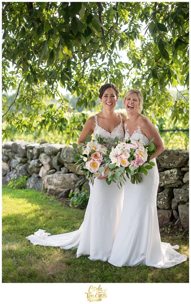 Two brides laugh by a stone wall at their Jonathan Edwards Winery Wedding