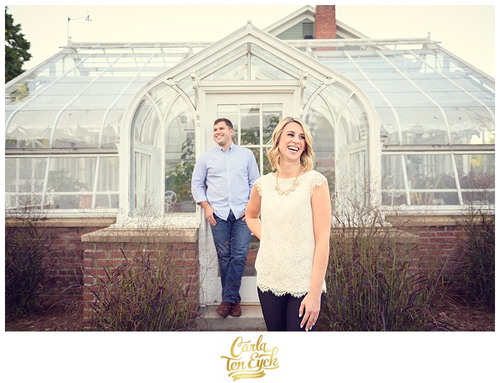 A couple laughs in front of a classic greenhouse in Elizabeth Park during their CT engagement session in Hartford CT