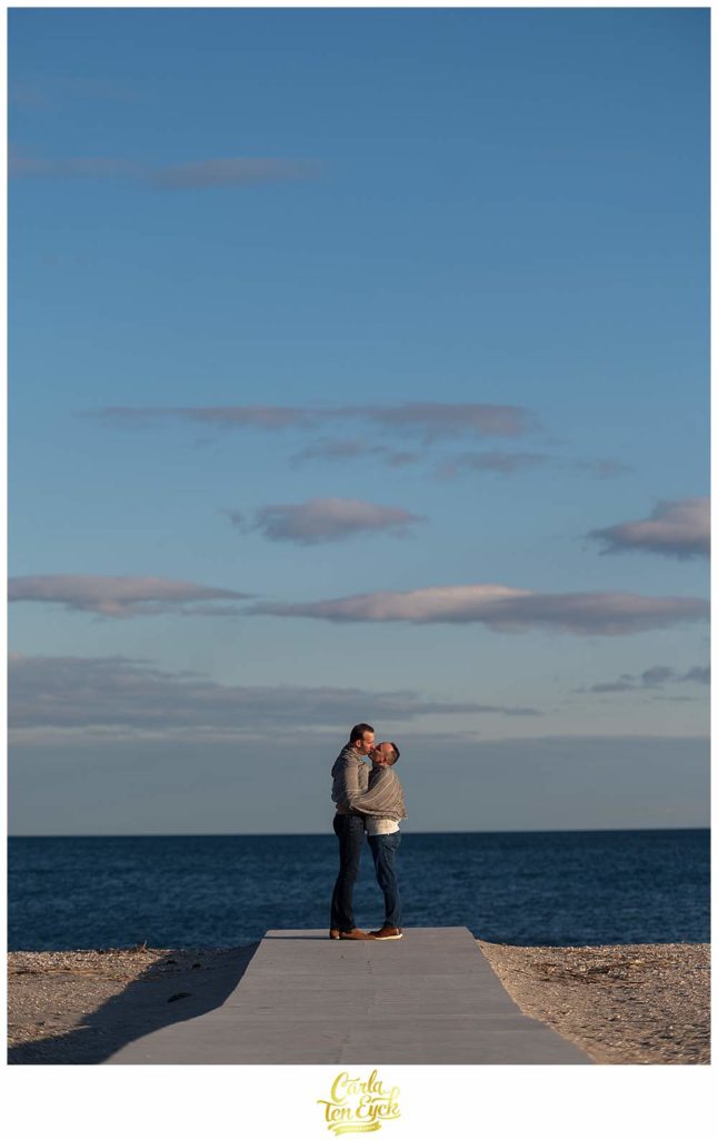 Two grooms kiss on the boardwalk during their winter beach engagement session at Silver Sands in Milford CT