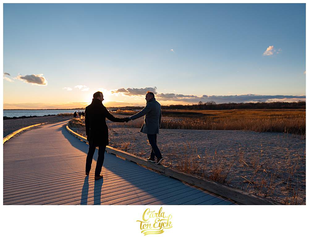 Two grooms walk at sunset during their winter engagement session at Silver Sands Beach in Milford CT