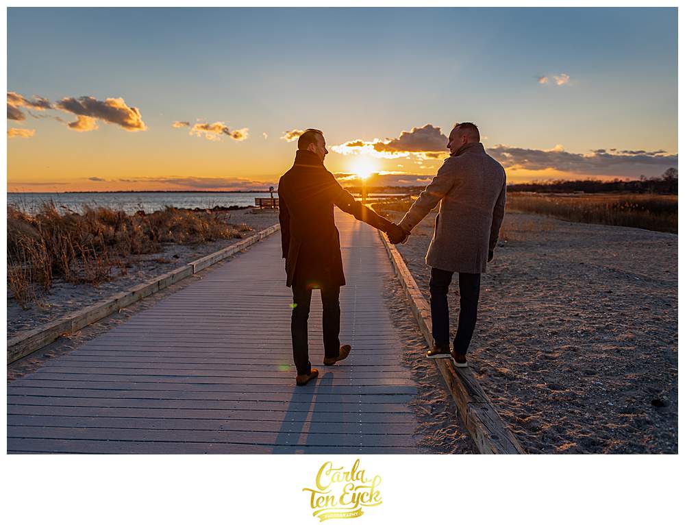Two grooms walk at sunset during their winter engagement session at Silver Sands Beach in Milford CT