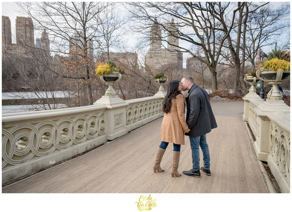 A couple kisses on the iconic Bow Bridge during their winter engagement session in Central Park NYC