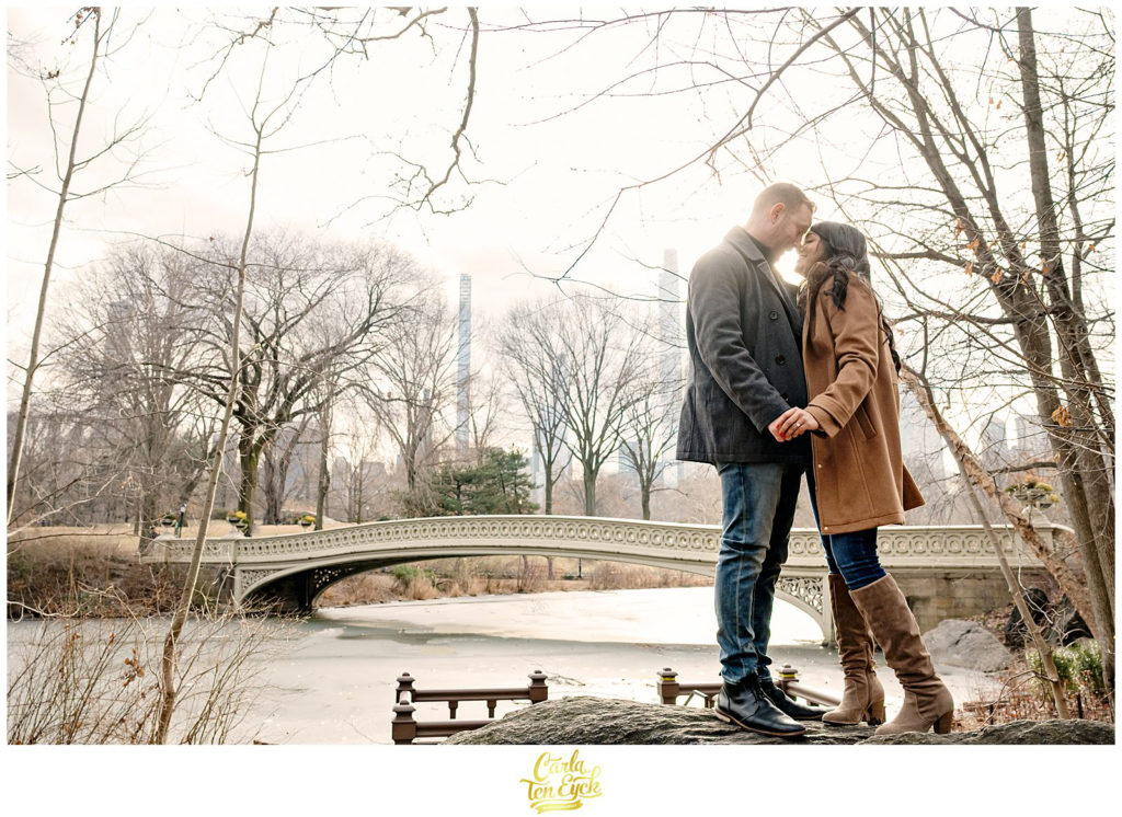 A couple kisses during their winter engagement session by the Bow Bridge in Central Park in NYC