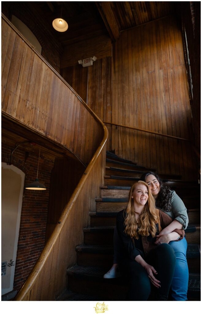 Two brides laugh while cuddling during their engagement session at  Mill NO. 5 in Lowell MA