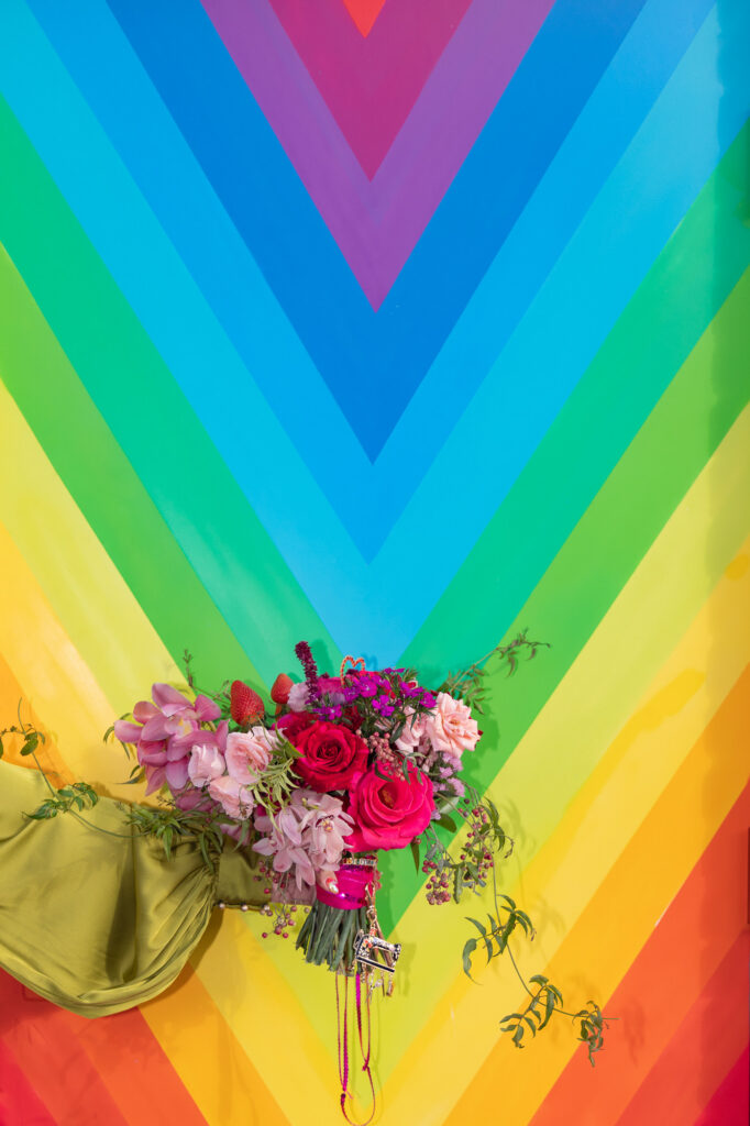 A queer bride holds her red and pink colorful wedding bouquet in front of a rain bow mural in Hartford Ct during her Hartford City Hall elopement