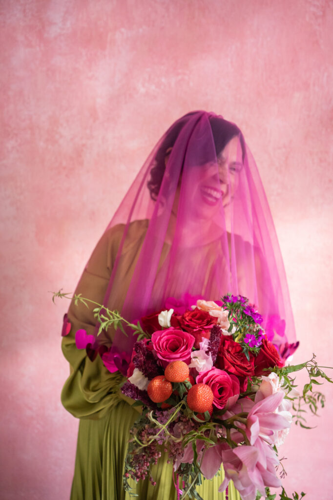 A queer bride laughs joyously at her colorful Hartford City Hall elopement in her hot pink veil holding her red and pink bouquet with strawberries